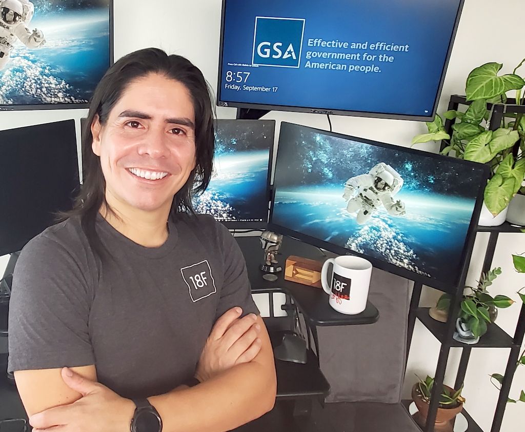 Edwin (el) posa sonriendo en frente de su escritorio. El tiene piel blanca bronzeada y largo cabello negro.  En su escritorio hay 5 monitores, uno se encuentra apagado, tres de ellos tienen la foto de un astronauta en el espacio, y el quinto el logo del GSA. En su escritorio tambien hay una taza de cafe con el logo de 18F, una decoracion de madera con la bandera de Puerto Rico, y un figurin del Mandalorian. A la par de su escritorio hay un estante con tres plantas verdes